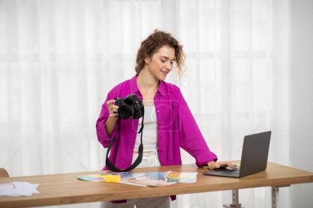 Téléchargez les photos : Femme photographe souriante tenant un appareil photo et utilisant un ordinateur portable tout en travaillant au bureau à domicile, jeune femme heureuse traitant des images sur l'ordinateur tout en se tenant au bureau près de la fenêtre - en image libre de droit
