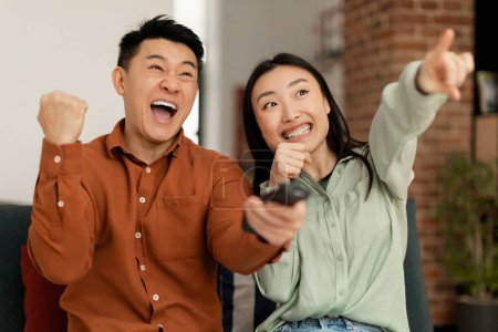 Photo for Joyful asian spouses shaking fists and shouting, watching sport channel, woman pointing on TV, sitting on sofa at home. Sport and television programming, weekend leisure - Royalty Free Image