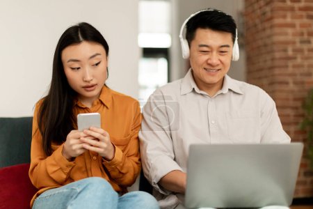 Téléchargez les photos : Jeune femme asiatique espionnage mari avec ordinateur portable, femme attrapé homme textos avec maîtresse, assis à côté de l'autre. Méfiance envers la famille et concept de trahison - en image libre de droit
