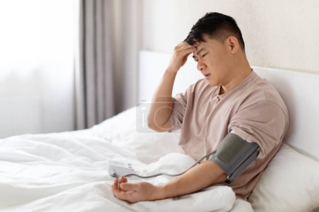Foto de Hombre asiático malsano usando pijamas sentados en la cama en casa, tocando la cabeza, sufriendo de dolor de cabeza, comprobando la presión arterial por su cuenta, usando un tonómetro moderno, espacio para copiar - Imagen libre de derechos