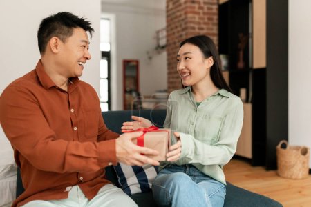 Photo for Loving asian man giving birthday gift to her lovely wife, celebrating at home, couple having marriage anniversary celebration, exchanging gifts, copy space - Royalty Free Image