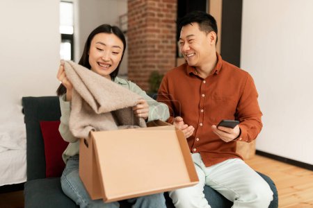 Foto de Emocionada pareja asiática desempacando caja de cartón en casa, compradores de hombre y mujer abriendo caja de cartón mientras están sentados en el sofá en la sala de estar. Compras en línea, pedido por Internet - Imagen libre de derechos
