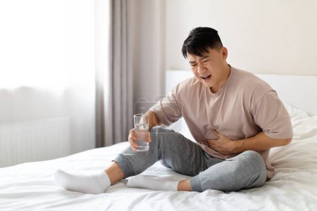 Photo for Unhappy sick middle aged asian man sitting on bed and touching his belly, suffering from stomach pain in the morning, wearing pajamas, holding glass of water. Gastrointestinal Diseases, Problems - Royalty Free Image