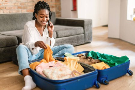 Téléchargez les photos : Heureuse femme noire assise près d'une valise ouverte avec des vêtements et parlant sur smartphone, réservant un voyage, faisant une réservation d'hôtel au téléphone depuis la maison, espace libre - en image libre de droit