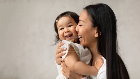 Téléchargez les photos : L'amour des mères. Joyeuse maman asiatique étreignant adorable petite fille bébé frottant le nez sur sa joue posant sur le mur gris à la maison. Maman câlinant avec le bébé. Happy Motherhood Moments. Panorama - en image libre de droit