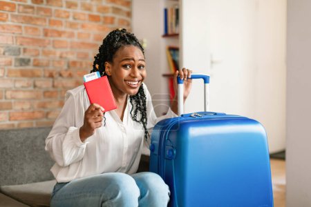 Foto de Mujer negra alegre sentada al lado de la maleta brillante, con pasaporte internacional con entradas y sonriendo a la cámara, lista para el viaje. Concepto de viaje de verano - Imagen libre de derechos