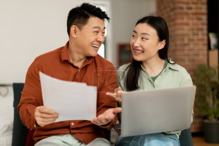 Téléchargez les photos : Couples mariés coréens joyeux payant leurs factures sur Internet via un ordinateur portable, assis ensemble sur le canapé, tenant des lettres et des papiers, utilisant l'ordinateur et souriant, profitant facilement e-banking - en image libre de droit