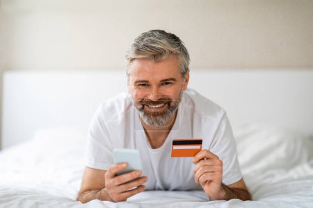 Foto de Feliz guapo de pelo gris barbudo hombre de mediana edad en ropa de casa acostado en la cama, sosteniendo el teléfono celular y la tarjeta bancaria, sonriendo a la banca de cámara en línea mientras se relaja en casa, espacio de copia - Imagen libre de derechos