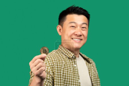 Foto de Retrato de hombre asiático feliz mostrando bitcoin y sonriente, enfoque selectivo en moneda de oro, fondo de estudio verde. Concepto criptomoneda - Imagen libre de derechos