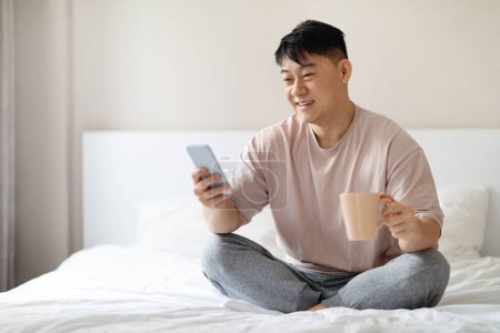 Photo for Cheerful happy positive handsome middle aged asian man wearing pajamas sitting on bed at home, using smartphone and drinking coffee, reading news, checking social media after waking up, copy space - Royalty Free Image