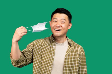 Photo for End of epidemic. Happy korean man taking off medical mask, feeling free and smiling over green studio background. Middle aged asian man removing facial protection, celebrating end of lockdown - Royalty Free Image
