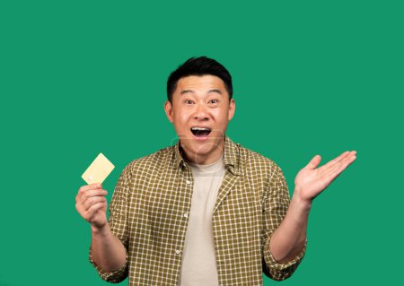 Téléchargez les photos : Excité homme asiatique d'âge moyen tenant la carte de crédit, levant la main vers le haut se réjouissant du succès isolé sur fond vert, plan studio. Gagnez, achats en ligne et cashback, annonce et offre - en image libre de droit