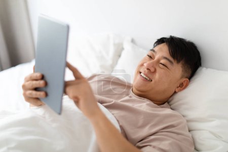 Téléchargez les photos : Positif heureux beau coréen d'âge moyen homme portant un pyjama en utilisant une tablette numérique moderne tout couché dans le lit le matin, vérifier les nouvelles en ligne, week-end de planification, en utilisant l'application mobile - en image libre de droit