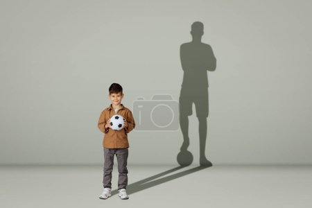 Téléchargez les photos : Heureux caucasien de 6 ans petit enfant décontracté avec ballon de football et l'ombre d'un joueur de football adulte isolé sur fond de studio gris. Avenir avec le sport, les rêves, le mode de vie enfant - en image libre de droit