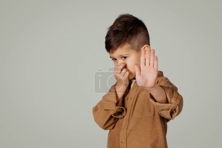 Photo for Sad caucasian 6 years old little boy covering nose with hand, making stop sign isolated on gray studio background. Stinks, no gesture, baby whims, kid emotions, disgust and facial expression - Royalty Free Image