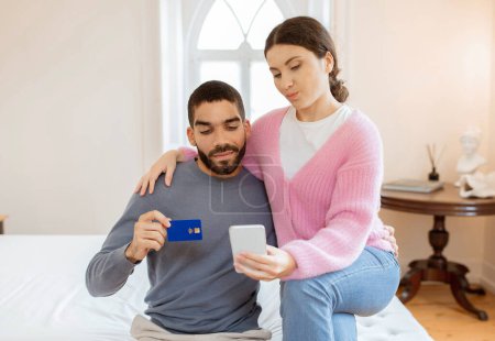 Photo for Couple Using Phone And Credit Card Shopping Online And Making Payment In Bank Application Sitting On Bed In Modern Bedroom At Home. Ecommerce Offer Concept. Selective Focus - Royalty Free Image