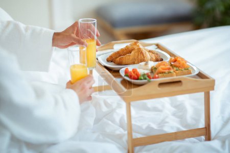 Photo for Unrecognizable Couple Having Romantic Breakfast In Bed Indoors, Holding Glasses With Orange Juice, Closeup Shot Of Table Tray With Delicious Food In Bedroom. Cropped, Selective Focus - Royalty Free Image