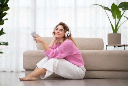 Portrait Of Happy Young Female Wearing Wireless Headphones Relaxing With Smartphone At Home, Cheerful Millennial Woman Listening Favorite Music While Resting On Floor In Living Room, Copy Space
