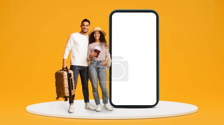 Photo for Glad young middle eastern family with travel tickets, suitcase near huge phone with blank screen on yellow studio background. Ready for vacation trip together, map app, trip blog, website - Royalty Free Image