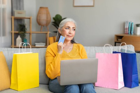 Photo for Pensive caucasian old female with a lot of bags with purchases sits on sofa enjoy shopping, thinks about order uses credit card and computer in living room interior. Sale and fashion blog at home - Royalty Free Image