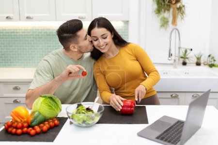 Photo for Loving handsome hispanic husband sitting at kitchen desk, helping his beautiful wife with cooking dinner, kissing her, holding cherry tomato, spouses watching movie on laptop and preparing meal - Royalty Free Image