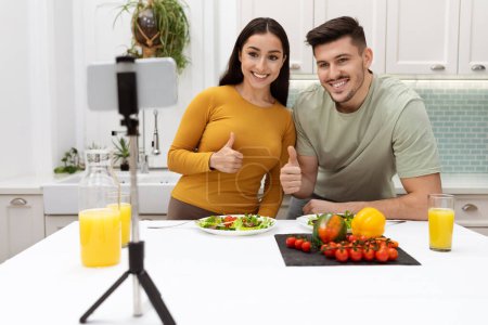 Téléchargez les photos : Beau couple heureux et aimant blogueurs gastronomiques en streaming pendant le dîner à la maison, homme et femme brune tirant contenu sur smartphone, montrant pouces levés, intérieur de la cuisine moderne, espace de copie - en image libre de droit