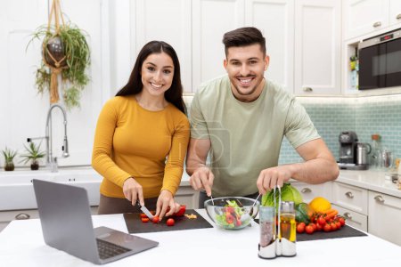 Téléchargez les photos : Portrait de heureux joyeux jeune couple attrayant portant occasionnel profiter du temps ensemble à la maison, cuisine saine délicieux dîner, couper des légumes frais, vérifier la recette en ligne, à l'aide d'un ordinateur portable - en image libre de droit