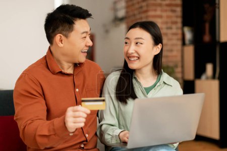 Photo for E-commerce, internet banking. Happy asian couple shopping online with laptop computer, sitting on couch at home and smiling at each other. Husband and wife making payment together - Royalty Free Image