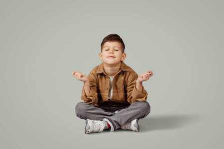 Foto de Feliz niño caucásico de 6 años en casual sentarse en el suelo con espacio libre aislado en fondo gris estudio. Meditar, emociones infantiles, estilo de vida infantil, descansar y relajarse - Imagen libre de derechos