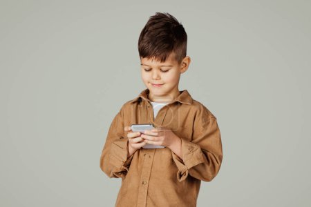 Photo for Smiling caucasian 6 years old little boy playing on phone game isolated on gray studio background. App for study, fun at free time with device, childhood and online education, gadget addiction - Royalty Free Image