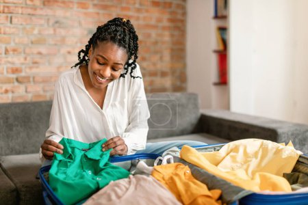 Photo for Suitcase preparation for vacation trip. Happy african american woman checking and packing clothes and stuff in luggage at home before travel - Royalty Free Image