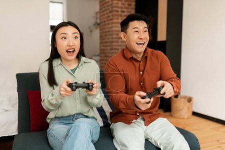 Téléchargez les photos : Joyeux conjoints asiatiques assis sur le canapé dans le salon et profiter de jouer à des jeux vidéo, passer du temps ensemble à la maison. Couple heureux profiter du moment le week-end - en image libre de droit
