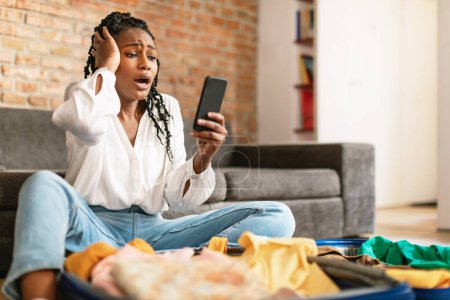 Téléchargez les photos : Femme afro-américaine terrifiée regardant l'écran du smartphone dans la terreur, se sentant choquée par l'annulation du vol, assise près de la valise ouverte à la maison, espace libre - en image libre de droit