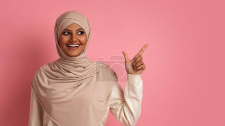 Photo for Nice Offer. Smiling Arab Woman In Hijab Pointing Aside At Copy Space With Finger While Standing Over Pink Studio Background, Happy Muslim Female Demonstrating Free Place For Advertisement - Royalty Free Image