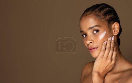 Foto de Retrato de mujer negra de mediana edad aplicando crema facial antienvejecimiento o hidratante, posando sobre fondo de estudio marrón, espacio para copiar. Cuidado de la cara, elevación, anti-envejecimiento, concepto de cosméticos nutritivos - Imagen libre de derechos