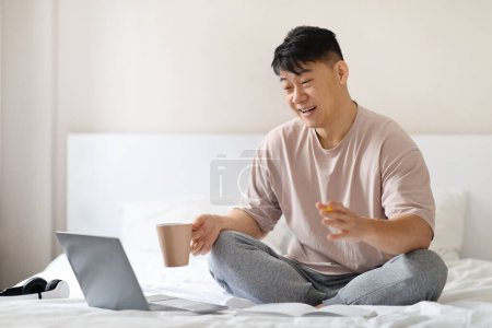Téléchargez les photos : Joyeux bel homme d'âge moyen portant un pyjama assis sur le lit à la maison ou à l'hôtel, à l'aide d'un ordinateur portable, tenant une tasse à café, regardant l'écran de l'ordinateur, souriant et gestuel, avoir un chat vidéo, copier l'espace - en image libre de droit