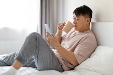 Photo for Relaxed happy handsome middle aged chinese man chilling in bed at home, using cell phone, drinking coffee, wearing pajamas, reading news online, checking email, side view, copy space - Royalty Free Image