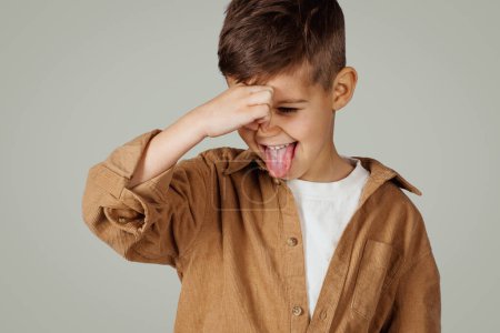Photo for Disgruntled caucasian 6 years old little boy in casual covering his nose with hand isolated on gray studio background, close up. Stinks, baby whims, child emotions, disgust and facial expression - Royalty Free Image
