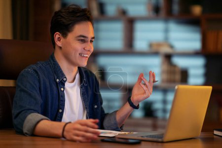 Photo for Joyful young businessman sitting at desk looking at computer screen talking with business partners, make informal video call, worker have online meeting with colleagues, working late at dark office - Royalty Free Image