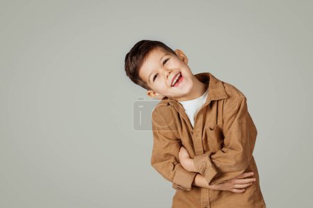 Téléchargez les photos : Riant mignon caucasien 6 ans petit enfant en presses occasionnelles mains au ventre isolé sur fond de studio gris. Mode de vie, enfance, éducation, plaisir seul, publicité et offre - en image libre de droit