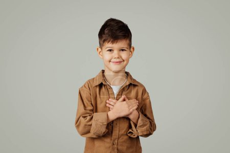 Photo for Cheerful caucasian 6 years old little boy in casual presses hands to heart, say thanks isolated on gray studio background. Grateful sign, child emotions, childhood and facial expression - Royalty Free Image