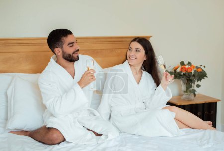 Téléchargez les photos : Couple romantique multiethnique buvant du vin mousseux ayant un rendez-vous romantique à l'hôtel. Conjoints appréciant Champagne assis sur le lit dans une suite de luxe en vacances de lune de miel, portant des peignoirs blancs - en image libre de droit