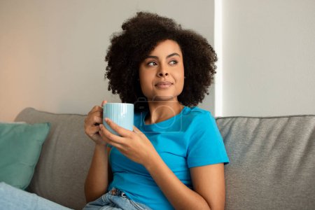 Téléchargez les photos : Sournois souriant millénariste afro-américain bouclé femme profiter de détente et une tasse de thé, regarder l'espace libre sur le canapé dans le salon minimaliste intérieur. Temps libre, soirée détente seule - en image libre de droit
