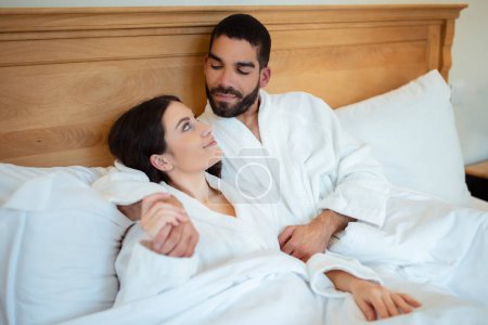 Photo for Diverse Couple In Love Embracing Lying In Bed In Bedroom Indoors, Wearing White Bathrobes. Spouses Looking At Each Other Relaxing At Hotel Suite On Vacation. Romantic Honeymoon - Royalty Free Image