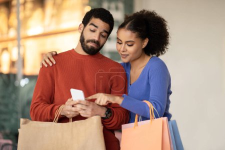 Photo for Mobile Shopping Offer. Diverse Buyers Couple Using Smartphone Choosing Clothes, Wife Pointing Finger Recommending New Mobile Application Standing At Modern Mall On Weekend. Ecommerce - Royalty Free Image