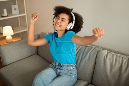 Glad millennial african american curly female in wireless headphones listen music, dancing, enjoy free time on sofa in living room interior. Fun and entertainment alone, audio app at home