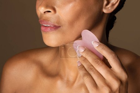 Cosmetic natural trend. African american woman using beauty Gua Sha quartz stone for massage, standing on brown background, closeup. Traditional medicine and cosmetology concept.