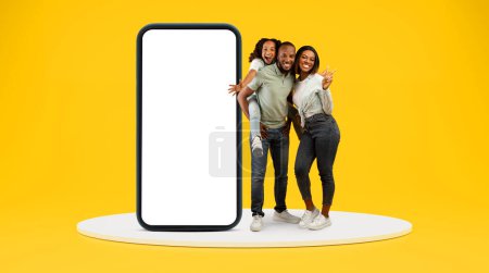 Photo for Happy african american family with small daughter have fun together with big phone with empty screen on stage on yellow background, studio. Parents and child, app, full length - Royalty Free Image