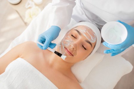 Photo for Skincare Treatment. Cosmetologist Applying Moisturising Mask On Face Of Beautiful Young Woman, Relaxed Attractive Female Enjoying Beauty Procedures At Luxury Spa Salon, Top View Shot - Royalty Free Image