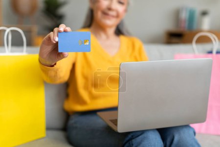 Photo for Glad caucasian retired woman with many bags from store sits on sofa, enjoy shopping, recommends credit card, use computer in living room interior, cropped. Home order, sale, banking and check finance - Royalty Free Image